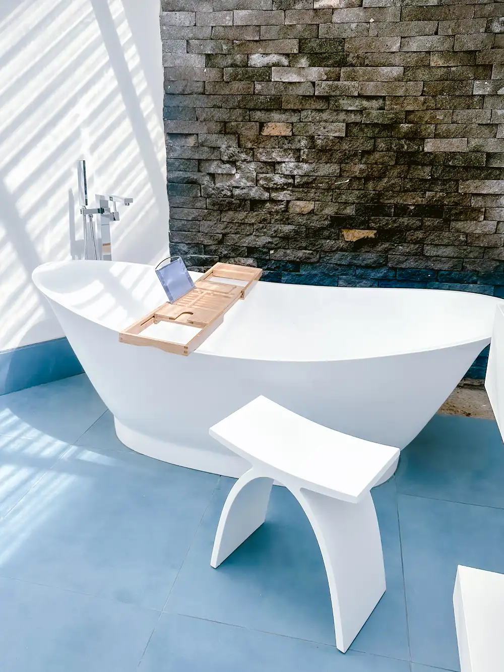 White oval freestanding bathtub with a wooden tray, on blue flooring and in front of a textured stone wall.